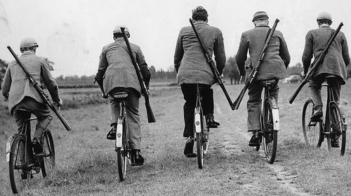 Men ride bicycles while carrying rifles.