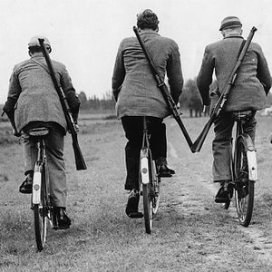 Men ride bicycles while carrying rifles.