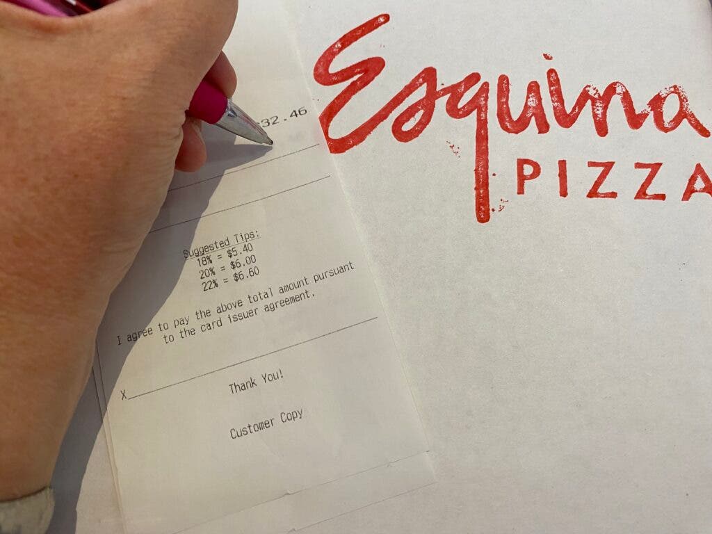 Someone debating how much to tip on a receipt atop a pizza box; options include 18, 20, and 22 percent.