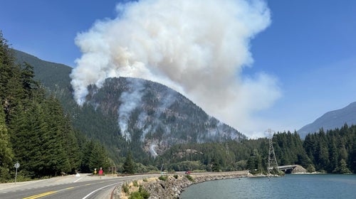 Wildfire smoke rises from a hillside next to a road running along a lake