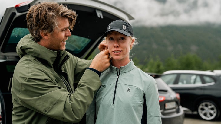 A female runner with a black cap on gets ready to run, while a man in a jacket fixes something to her ear.