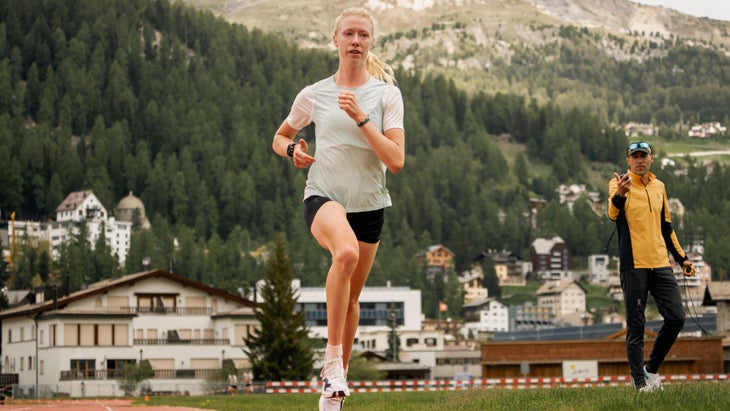 A woman runs around the track while a man in the background, her coach, times her on a stopwatch