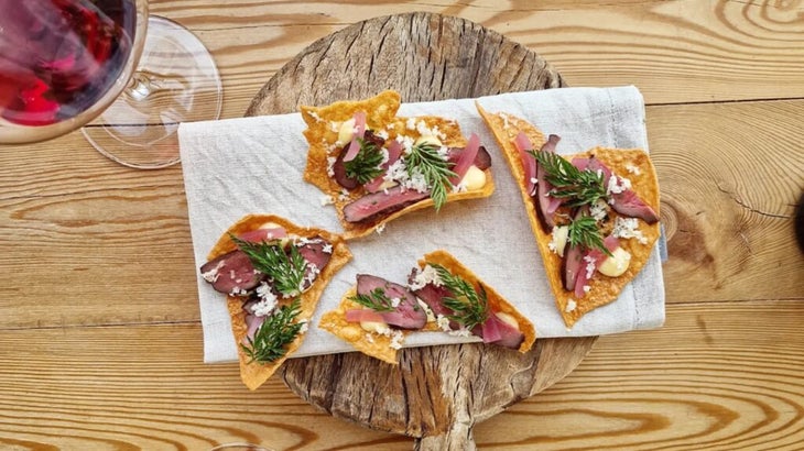 A circular wooden platter with crudités of grilled beef heart atop crackers of yellow peas, with a glass of red wine to the left