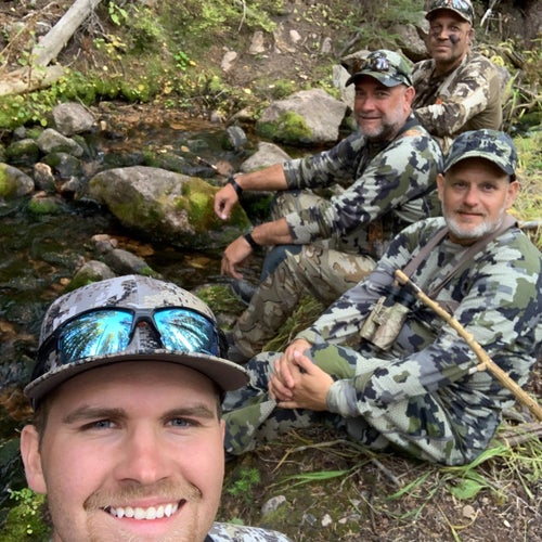 Four men in hunting gear smiling at the camera and sitting at a mountain stream