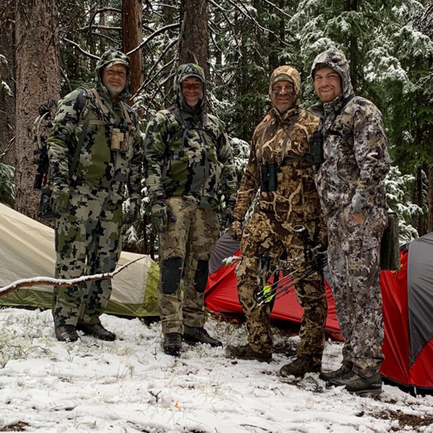 Four men in hunting gear stand in front of their tents in a forest, with snow on the ground