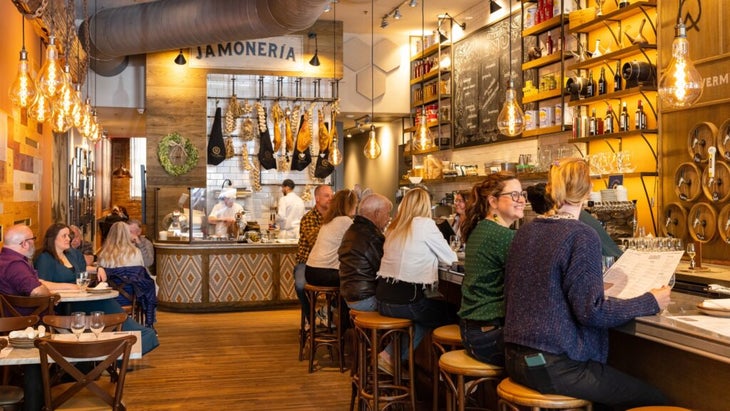 People sitting at tables and at the bar of Curate, a notable restaurant in Asheville. A sign in the back above hanging cured ham hocks, reads "Jamoneria."