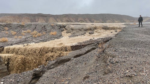 A person stands in a rocky desert landscape next to a newly formed river of muddy flood water.