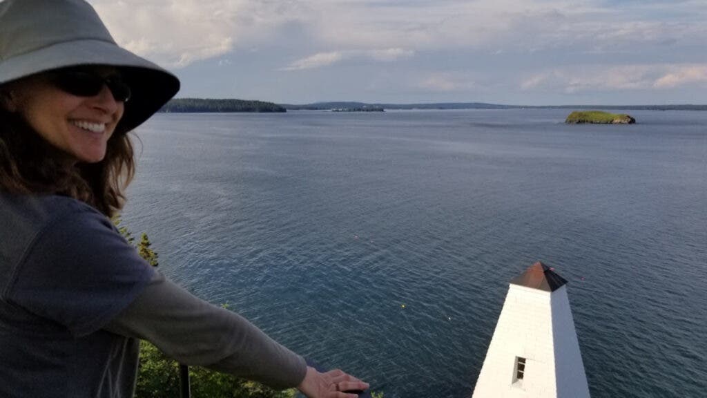 The author wearing a sunhat and dark sunglasses Eagle Island, Maine, with a lighthouse and Casco Bay spread out on the horizon