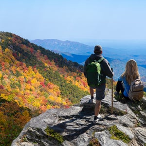 Two hikers—one woman, one man—look over a rock bluff at an expanse of Blue Ridge Mountains whose trees are bursting with yellow, red, and green leaves.