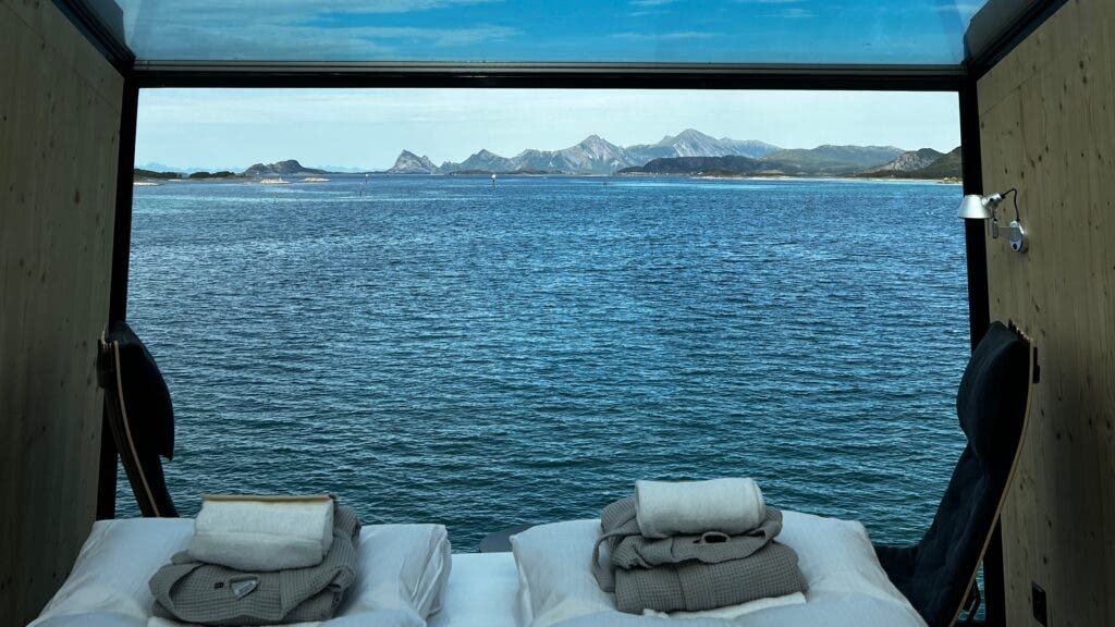 The interior of the second floor of the new towers, with a bed for two, two black chairs, and a glass ceiling. The view looks out at the sea and distant mountains.