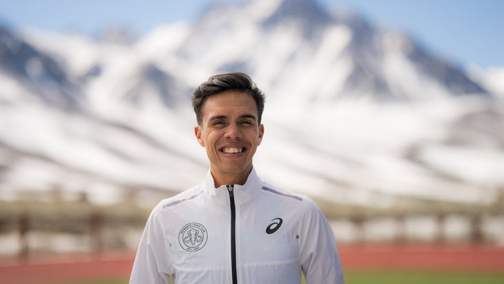 A man in a white windbreaker and black hair smiles in front of a mountainous backdrop