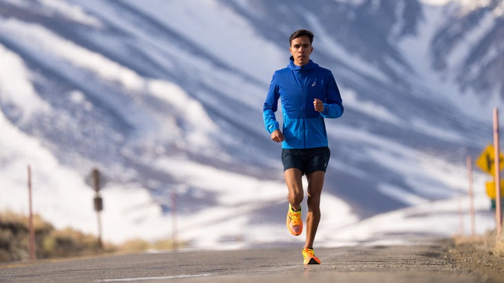 A man runs on a road with a blue jacket on and mountains in the background