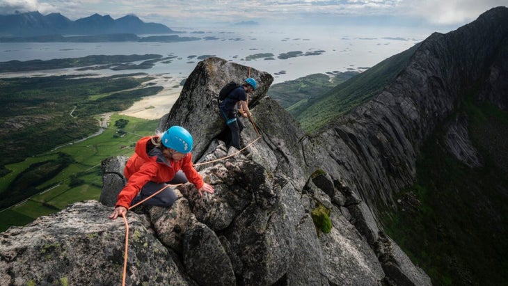 Two climbers navigate the precipice of the Nordskot Traverse, with an expansive vista of a gray sea and verdant valley below.