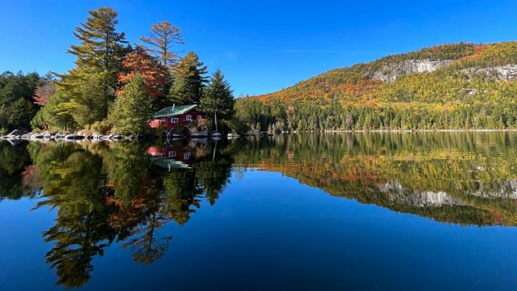 A red cabin set against hardwoods and a glassy lake reflecting a nearby mountain and fall foliage