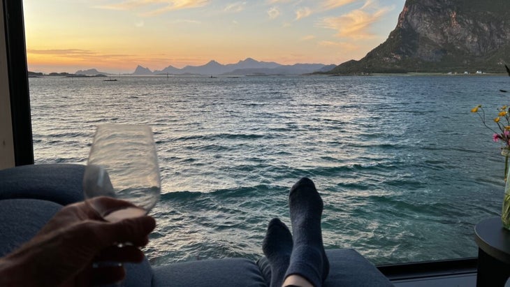 A pastel-colored sunset view over the waterfront and mountains. You can see a person relaxing with socks on, holding a glass of wine.