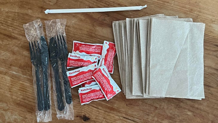 single-use take-out trash on table including napkins, ketchup packets, black plastic utensils in plastic and a straw