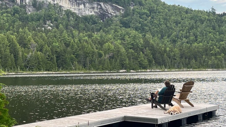A woman sits in an Adirondack chair on a wooden lake pier, with a white lab relaxing behind her