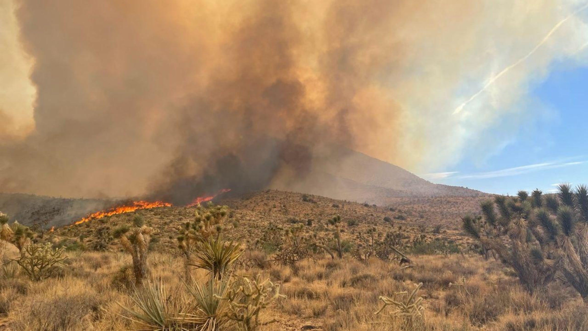 Fire and smoke billows over a hill formation in a desert landscape.