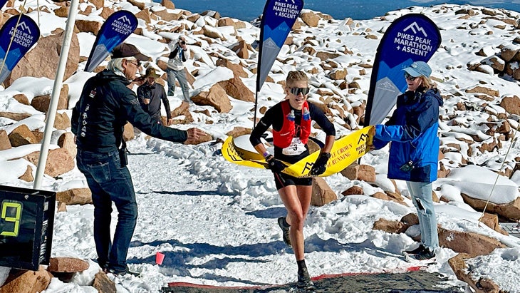a women in a red running vest breaks a yellow tape to win a race on the top of a mountain