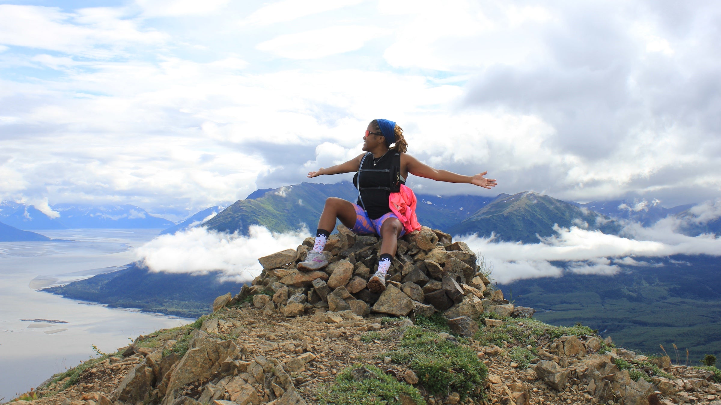 A woman in pink running bottoms spreads out her arms at the top of a mountain