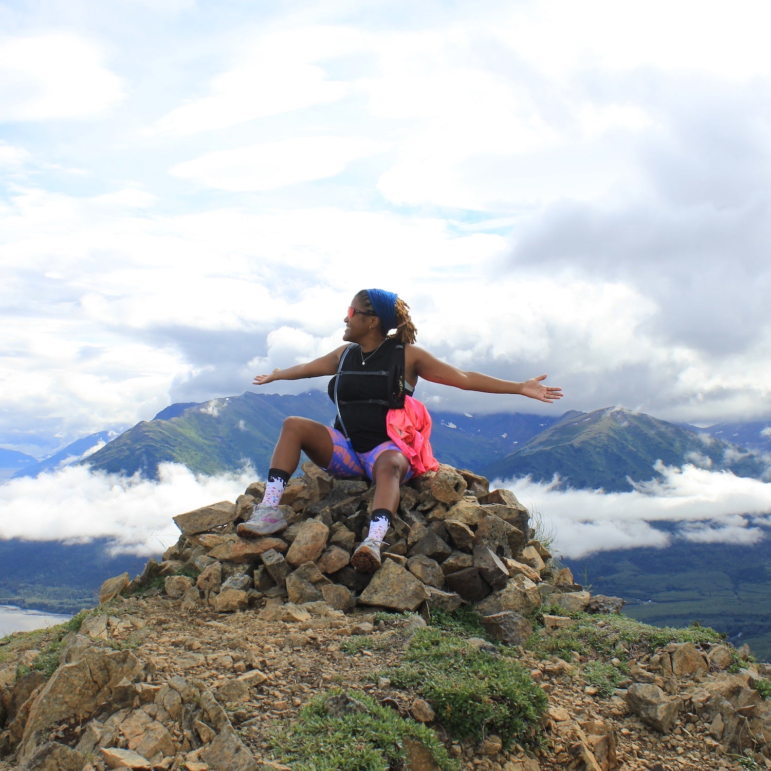 A woman in pink running bottoms spreads out her arms at the top of a mountain