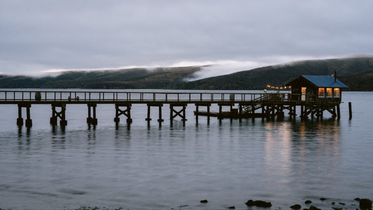 The boat shack at the end of a wooden pier at dusk, with the fog rolling over Point Reyes National Seashore in the background