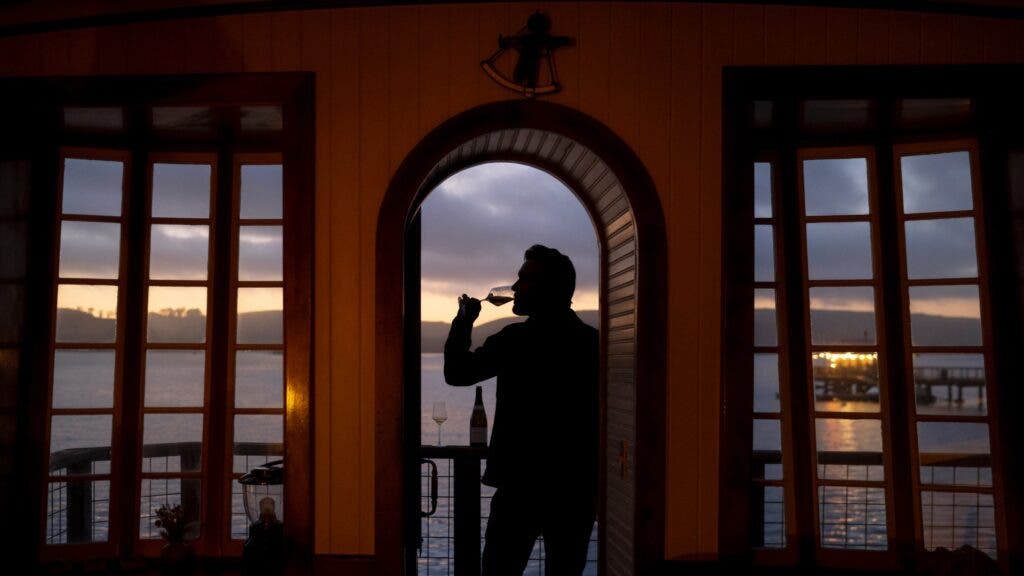 A man drinking a glass of wine in the doorway of one of the cottages, with Tamales Bay and the boat shack in the background