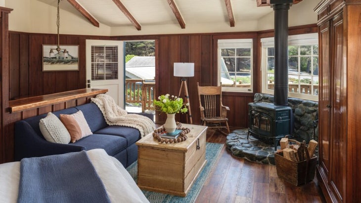 The main room of Jerry's cottage, with a sea-blue couch, wooden table with a vase of flowers, and a woodstock and rocking chair