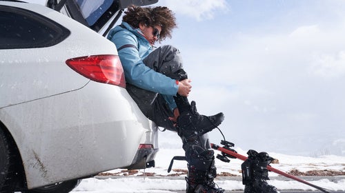 Man sitting in the trunk of a white car putting on ski boots.