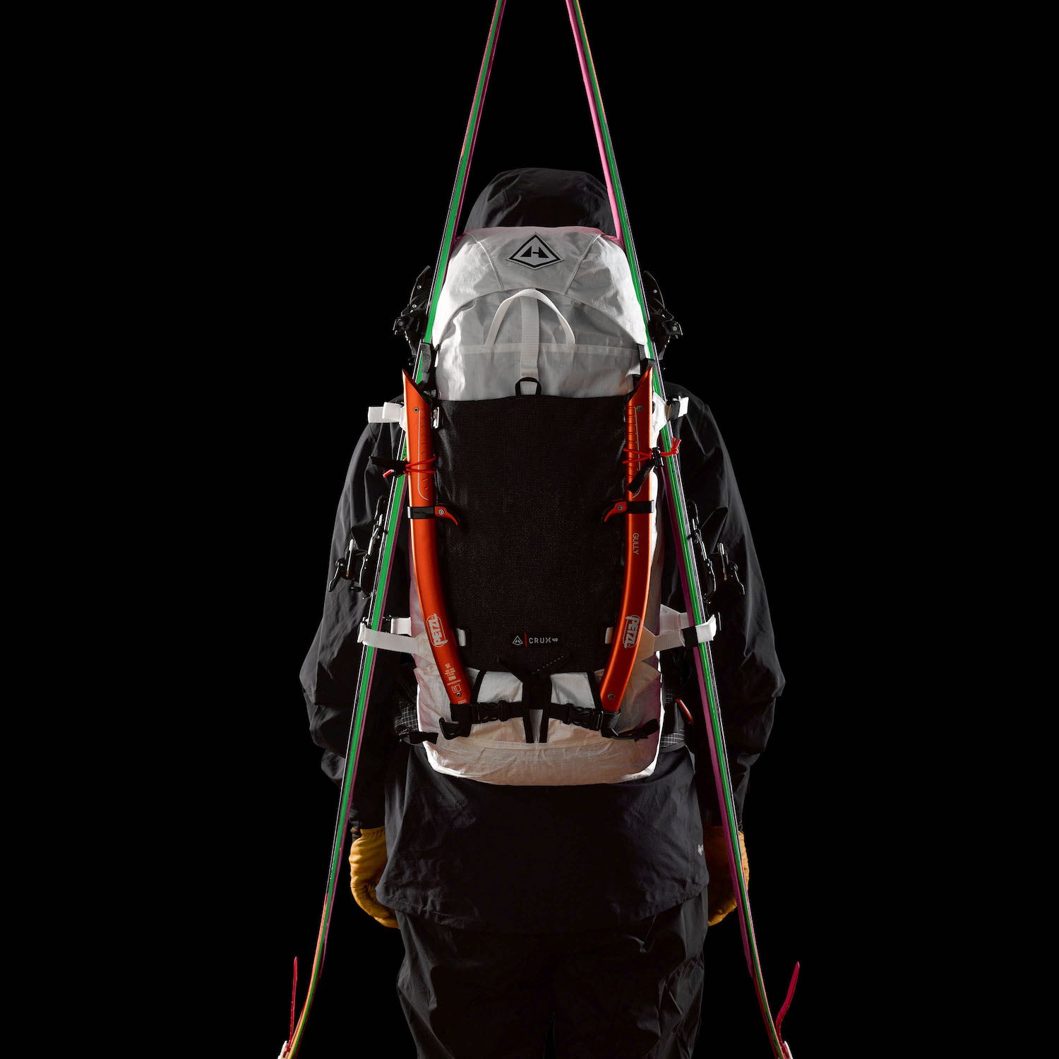 Person wearing white HMG Crux ski mountaineering pack with skis attached in front of black backdrop