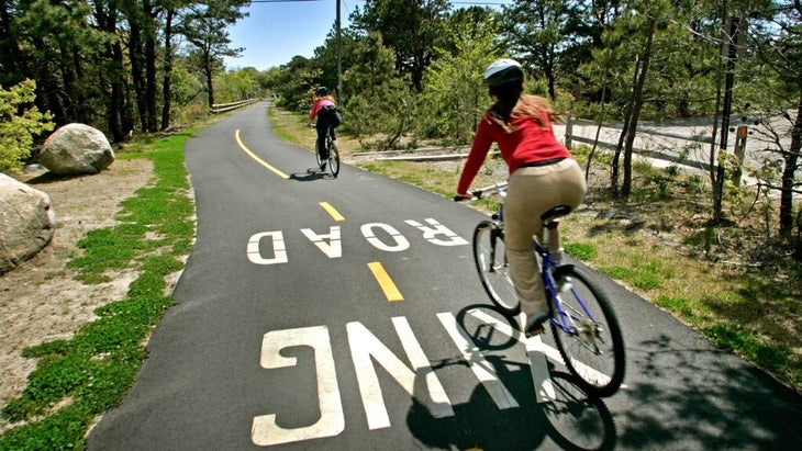 Two cyclists wearing helmets headed down the paved Cape Cod Rail Trail on a sunny day