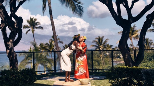 Two Hawaiian women, one wearing an orange muuuu and both with flowers in their hair, hug on an overlook surrounded by palm trees and the ocean in the distance.