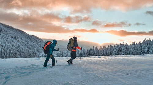 Two hikers dressed in warm winter sportswear with hiking backpacks walk with trekking poles in the snow-covered pine mountains in an incredible sunset, beautiful sky.
