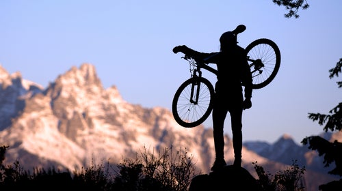 A silhouetted mountain biker holds his bike and admires the view from a ridge during an early morning ride through the Grand Teton mountains. The imaged is framed on all sides with foreground trees and the early morning sun on the Teton peaks in the background.