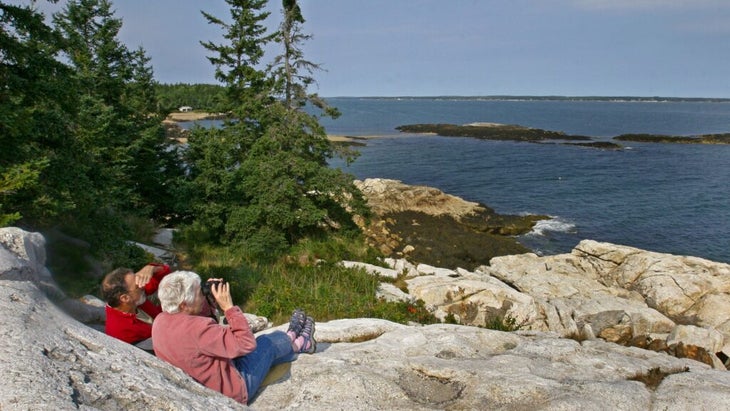 An older couple sitting together atop boulders, birding with a pair of binoculars