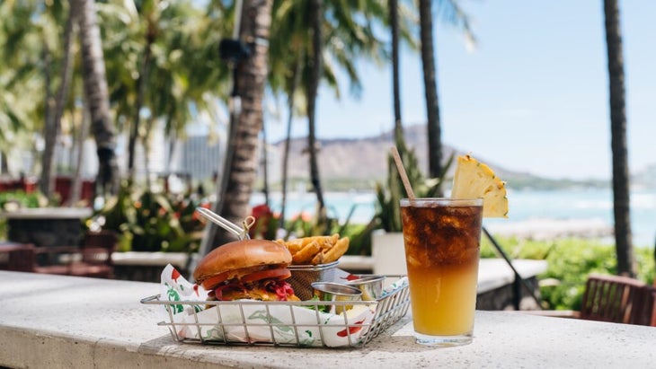 A burger and mai tai perched on a cement wall, with a background of Diamond Head, palm trees, and turquoise waters