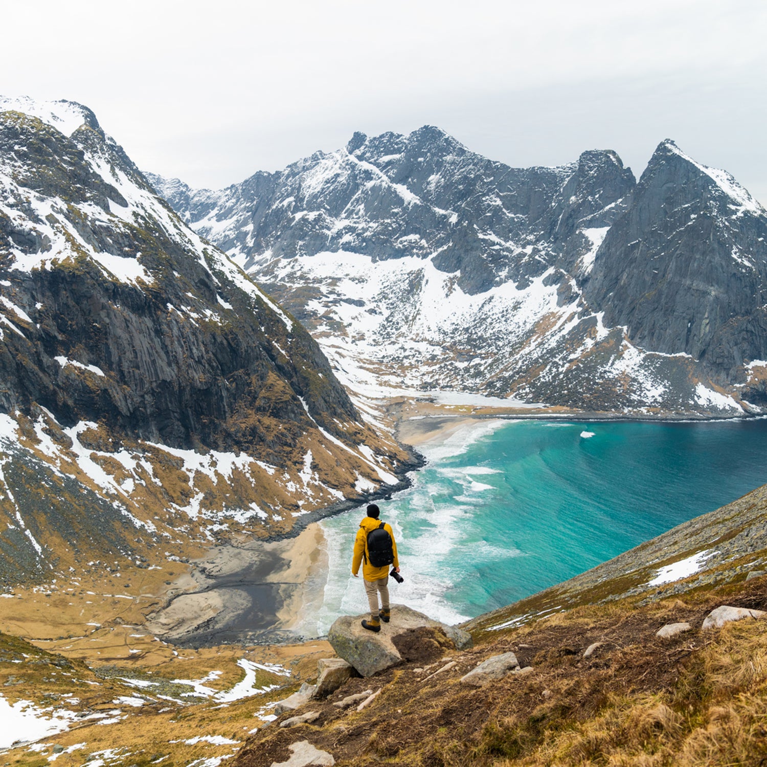 An unrecognizable person wearing a yellow jacket standing on a cliff looking down to a blue beach in Norway with high snowy mountains in the background.