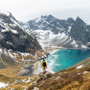 An unrecognizable person wearing a yellow jacket standing on a cliff looking down to a blue beach in Norway with high snowy mountains in the background.
