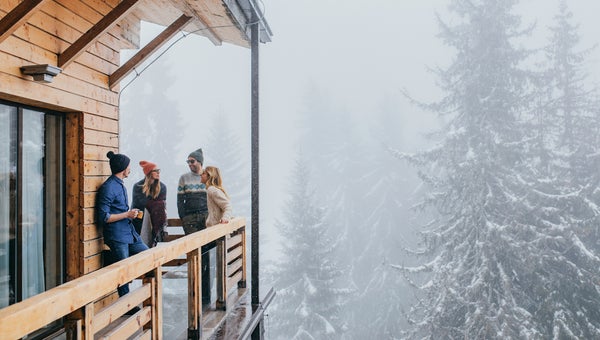 Group of men and women in winter outfits spending time on log cabin balcony in mountain.