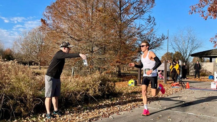 A man in a white shirt runs around a turn and picks up a bottle