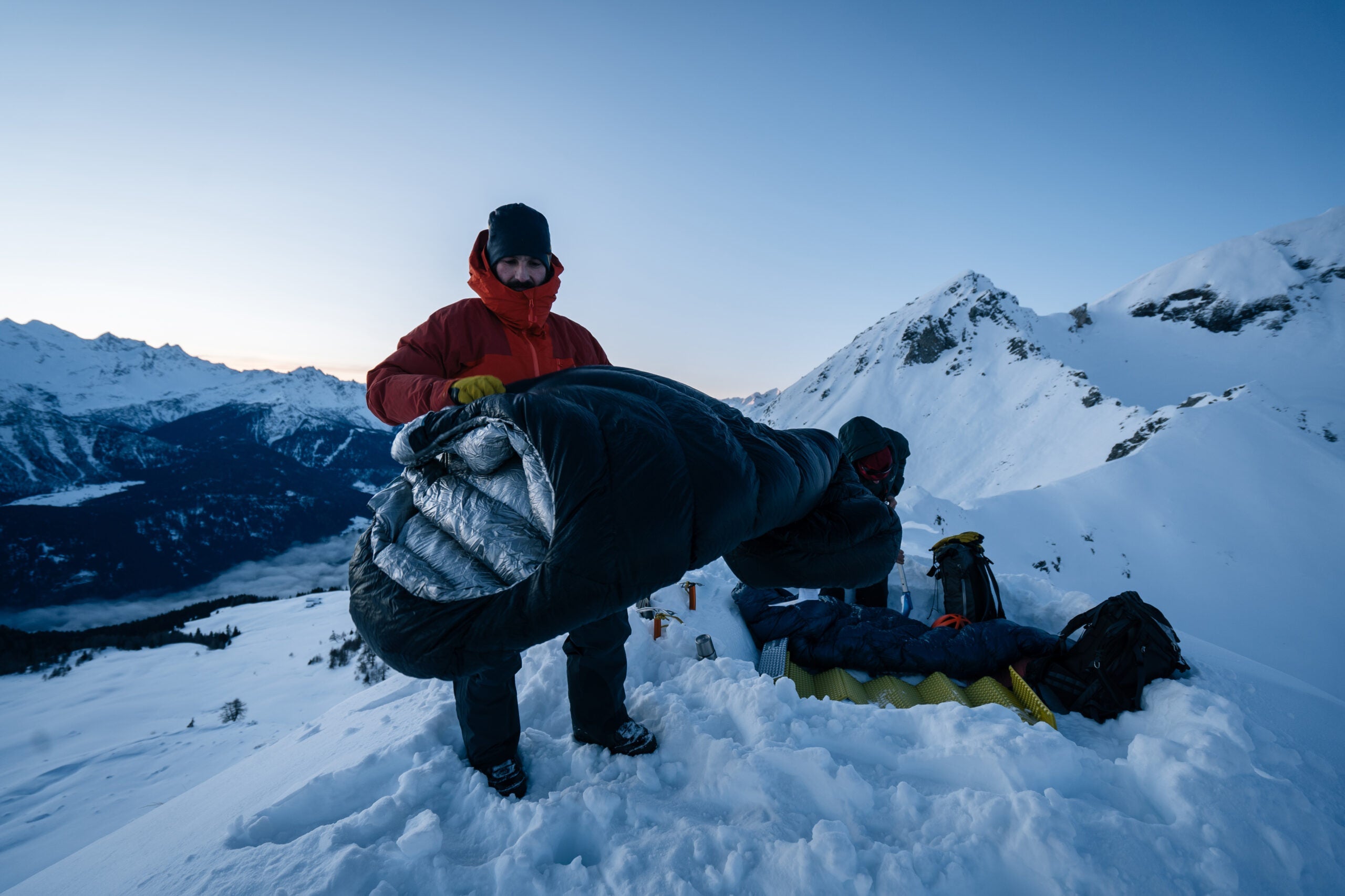 A man in cold weather gear holds a sleeping bag. He is standing on snow in front of snowy mountains and it is dusk.