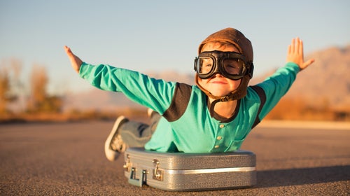A boy wearing aviator goggles pretends to fly; his arms are outstretched and he's lying prone atop a suitcase on a stretch of open road that looks like a runway.