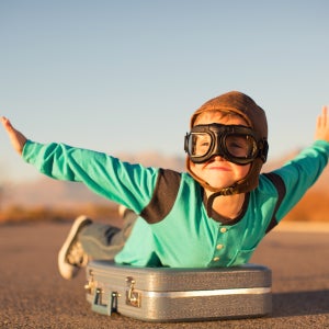 A boy wearing aviator goggles pretends to fly; his arms are outstretched and he's lying prone atop a suitcase on a stretch of open road that looks like a runway.