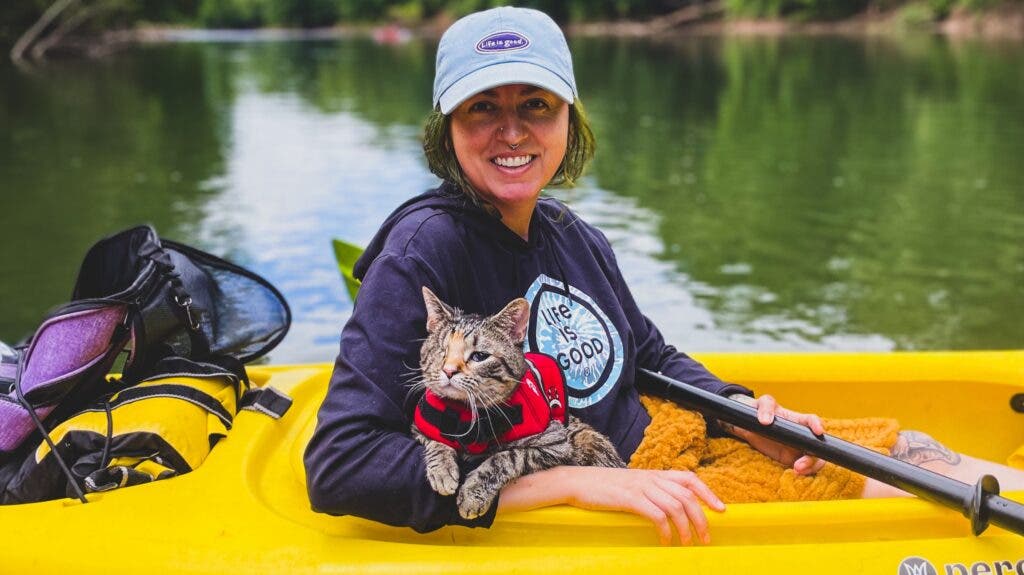 A woman a single kayak, with her one-eyed cat (wearing a PFD) in her lap, looking relaxed.