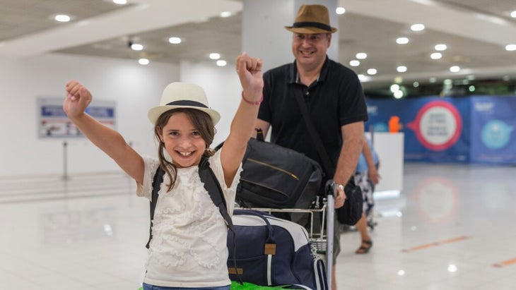 A gleeful girl with her hands raised in excitement sits on a luggage cart being pushed by her dad, who is wearing a hat and smiling.
