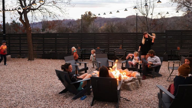 Families sitting around the fire pit at dusk, while children run about pebble-filled outdoor area that's fenced in