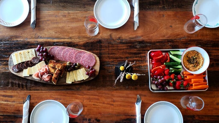 A table set with plates and silverware wrapped in knives, and to eat: a wooden platter of charcuterie next to a platter of cut fruits and vegetables