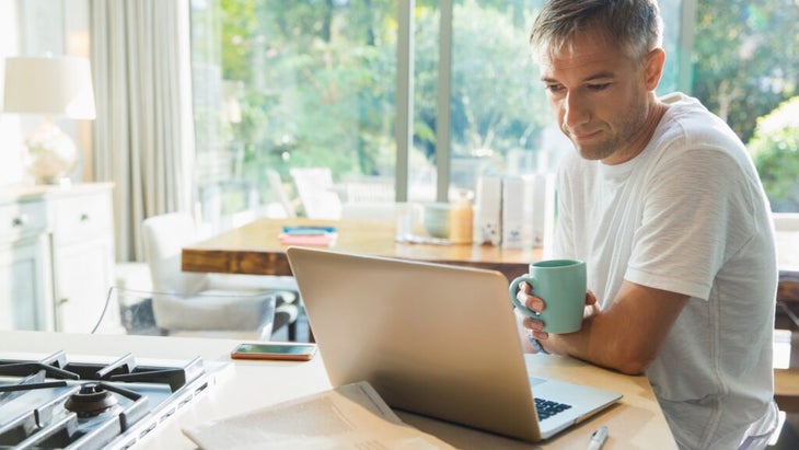 A man sitting at his laptop in the kitchen in the morning while holding a mug.