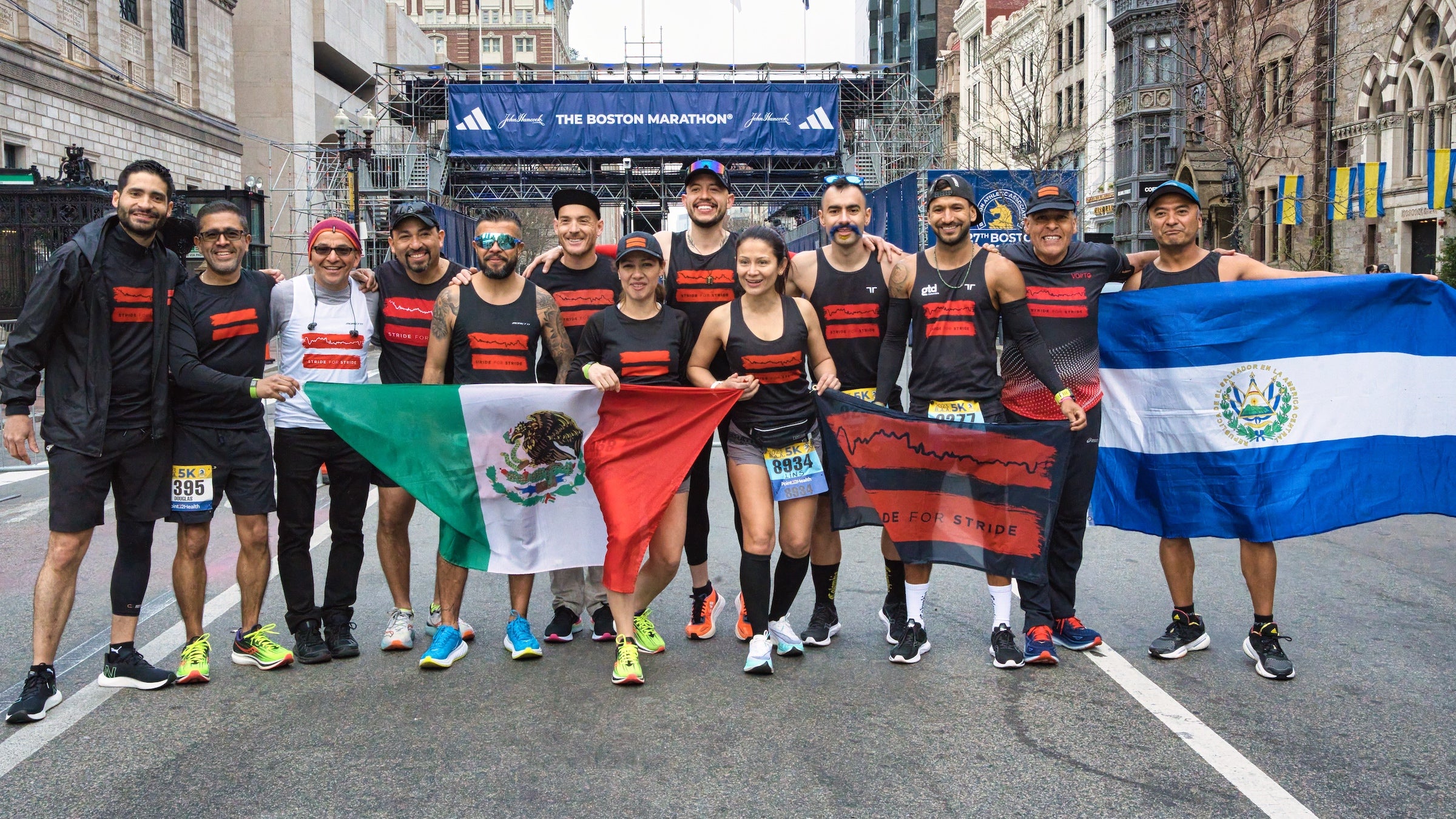 A group of runners with various flags stand in front of the Boston Marathon finish line.