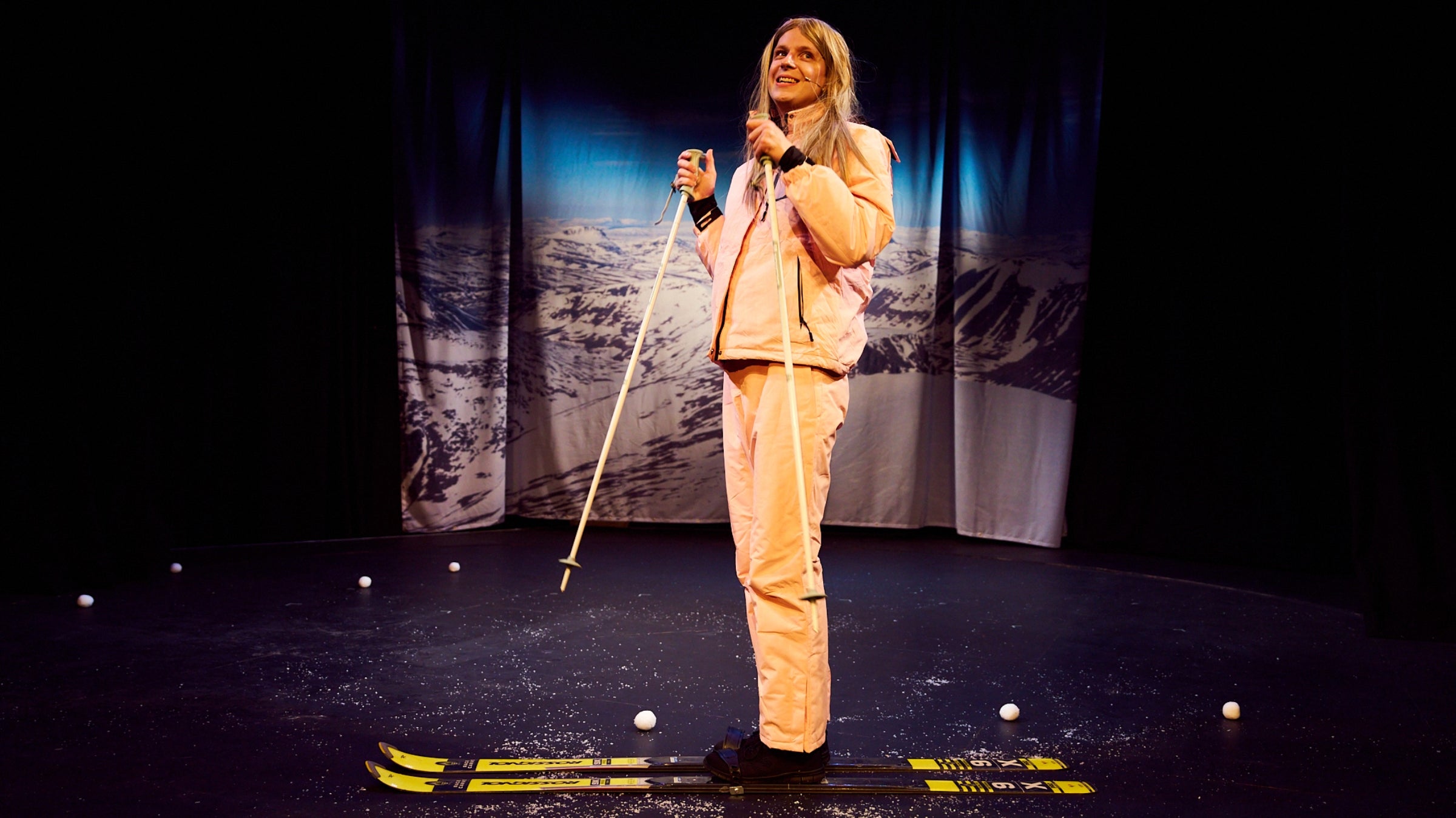 An actor in a long blonde wig stands on stage on a pair of skis